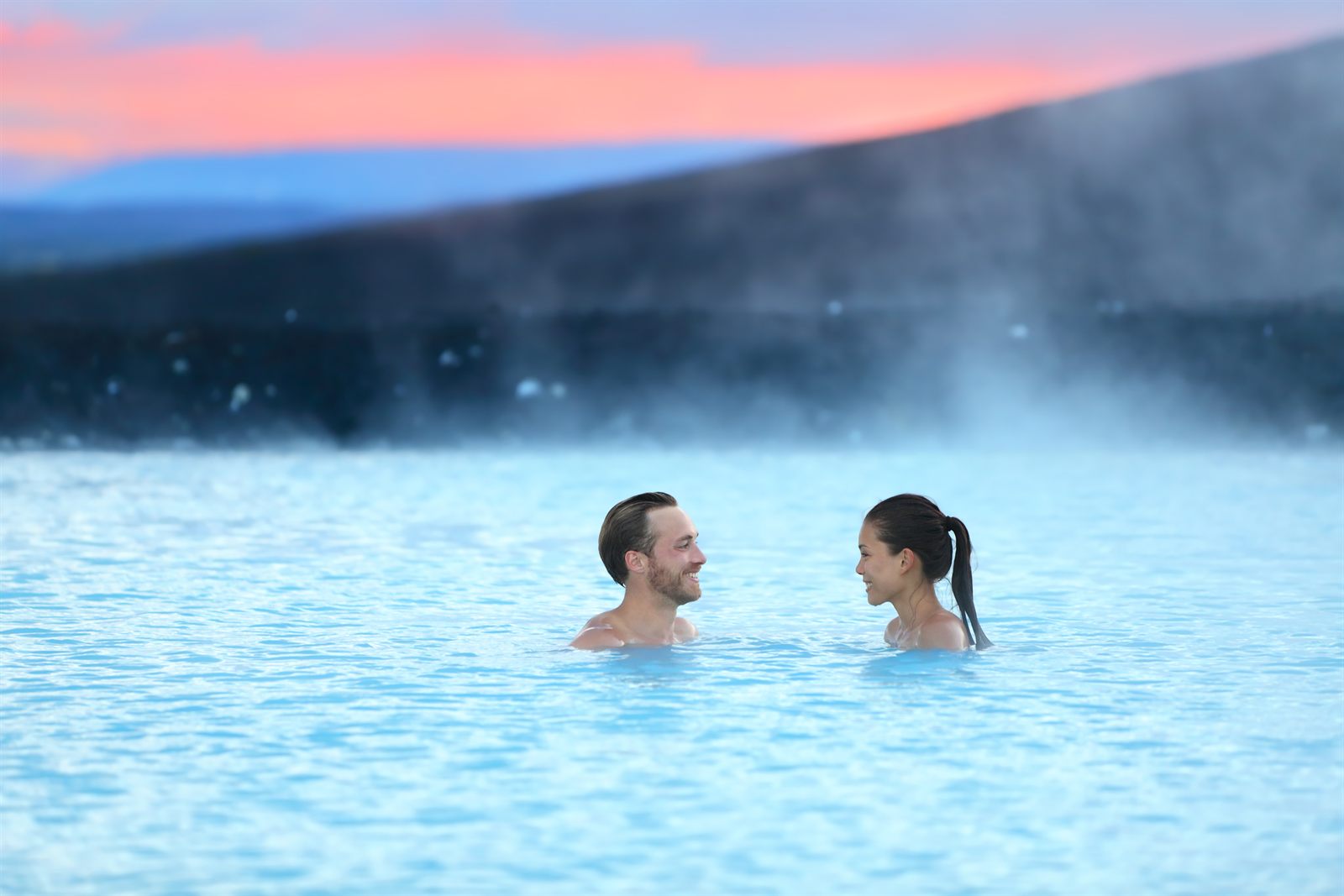 Couple relaxing in the steaming blue water of a geothermal lagoon at dusk, one of the travel agent perks of learning a destination