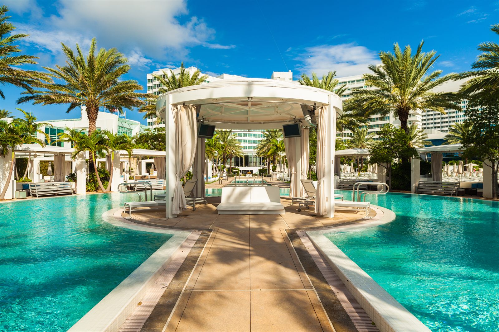 Resort swimming pool with a white cabana on a walkway between palm trees, the type of stay travel agent rewards programs can lead to